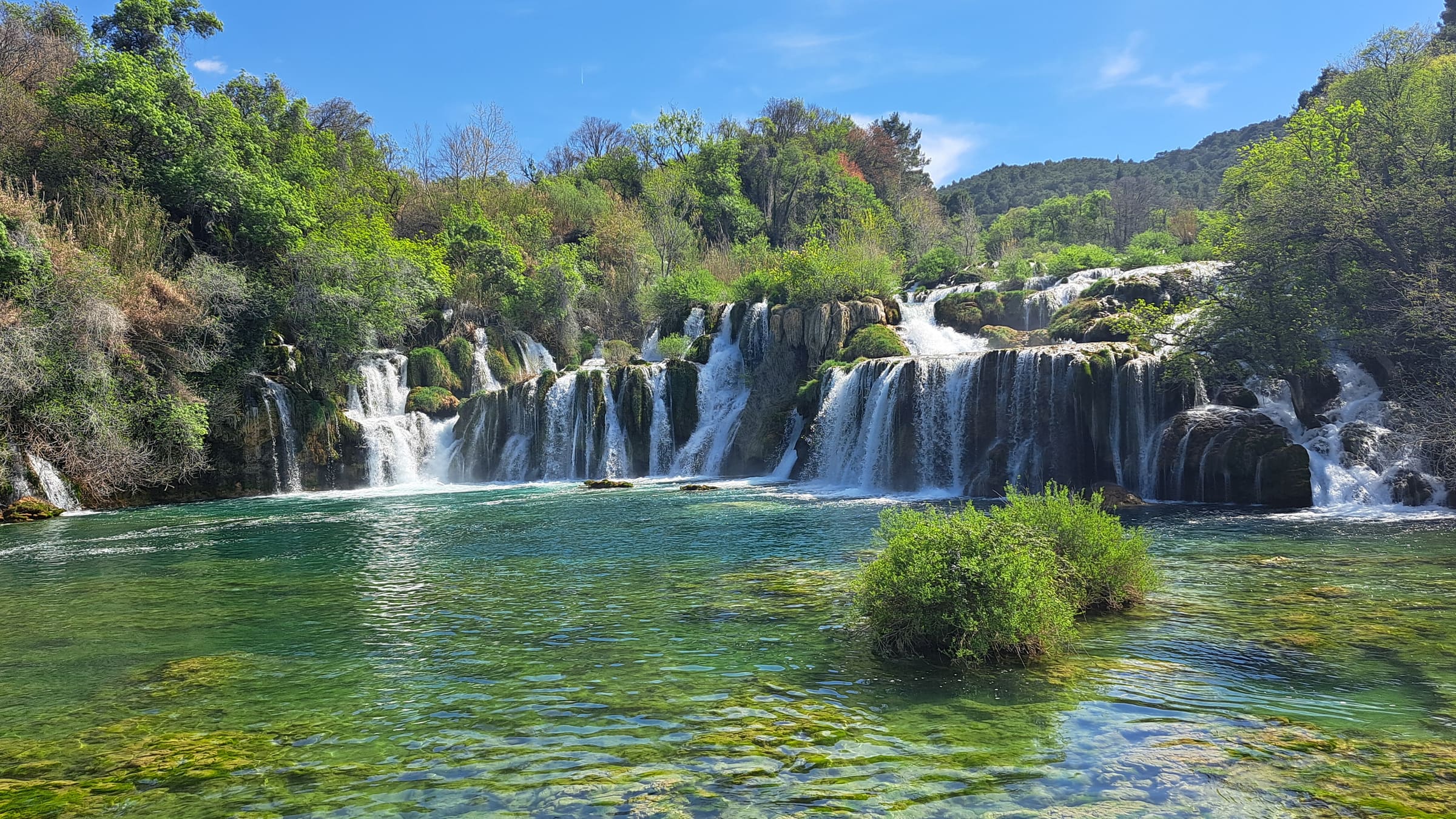Breite Wasserfallkaskade, die über bewachsene Felsstufen in ein türkisgrünes, flaches Becken fällt, umgeben von dichtem Grün unter blauem Himmel.