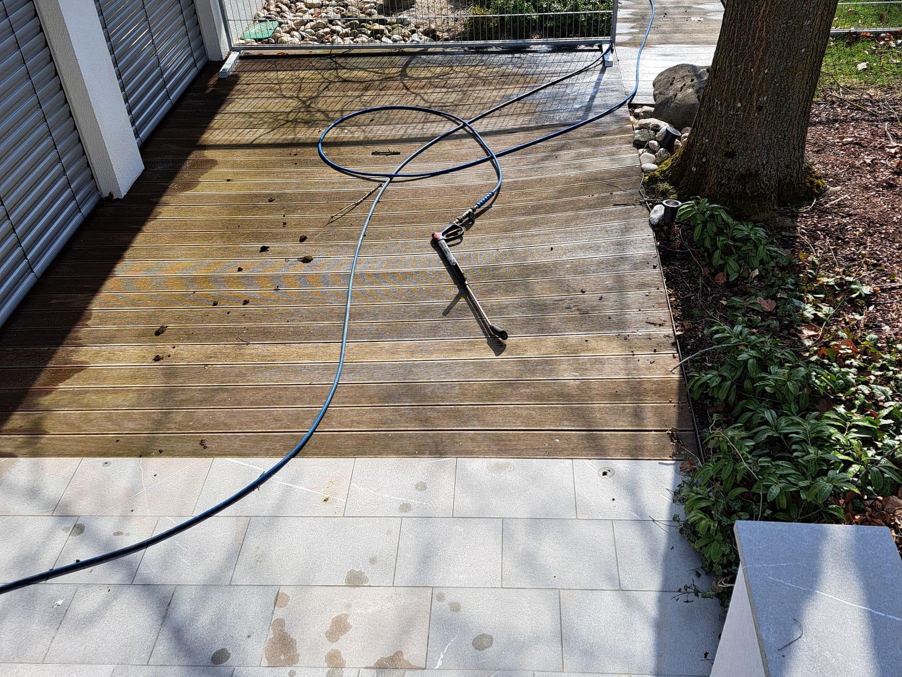 Terrasse mit Hartholzdeck und Steinplatten vor der Behandlung: vergrautes Holz und fleckiger Stein, Reinigungsschlauch im Bild.